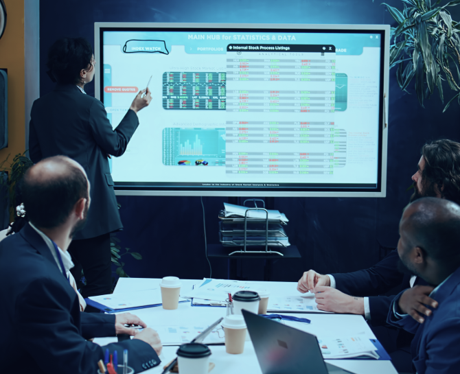 A professional meeting setting where a presenter points at a data-filled screen while attendees observe, surrounded by papers and coffee cups.