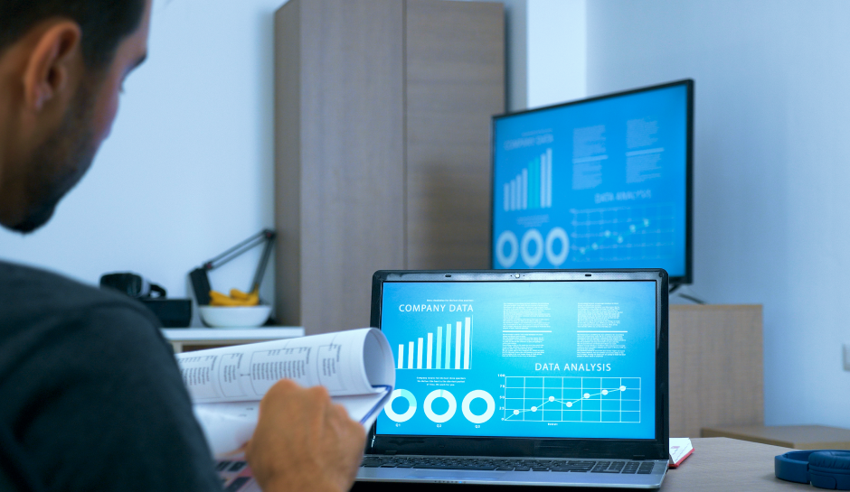 A person reviews data reports while looking at a laptop displaying graphs and charts in a modern workspace.