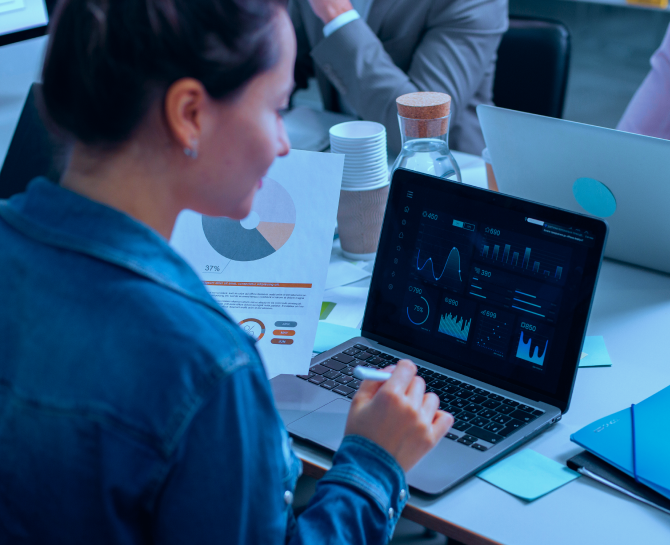 intent data in abm -A person in a denim jacket reviews data visualizations on a laptop, surrounded by papers and colleagues in a modern office setting.