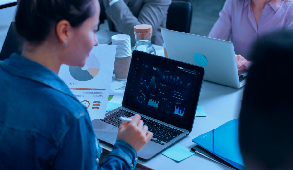 intent data in abm -A person in a denim jacket reviews data visualizations on a laptop, surrounded by papers and colleagues in a modern office setting.