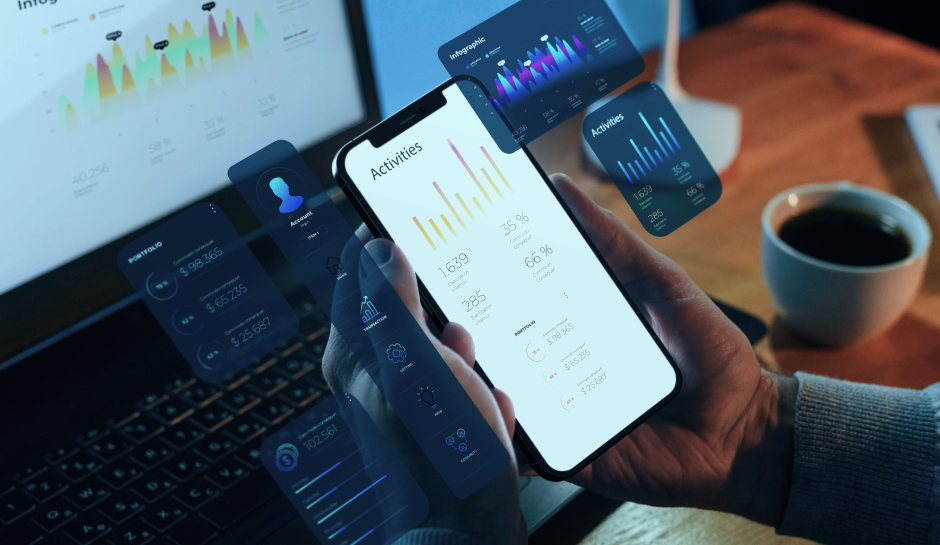 A person holds a smartphone displaying colorful graphs and activity metrics, with a laptop and coffee visible in the background.