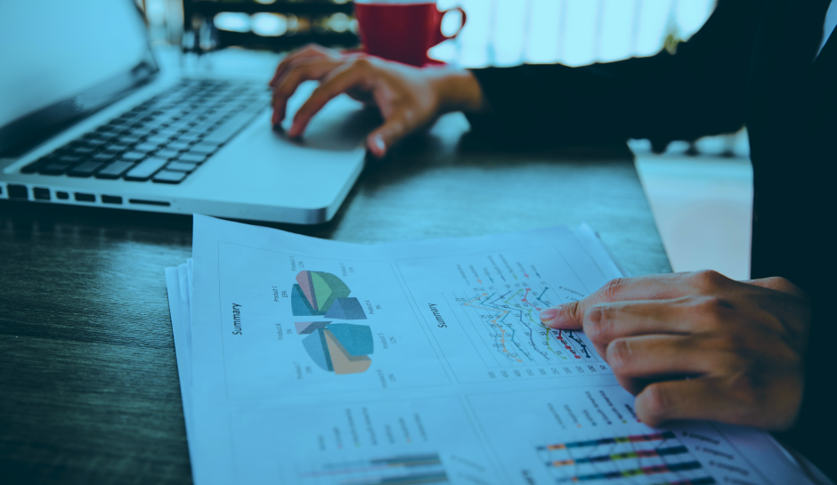 A person analyzing printed charts and graphs while using a laptop, with a red cup in the background on a wooden desk.