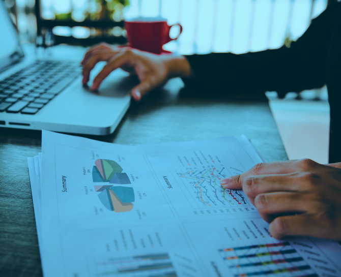 A person analyzing printed charts and graphs while using a laptop, with a red cup in the background on a wooden desk.