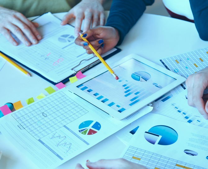 Hands of multiple people working together, analyzing charts and graphs on printed documents and a tablet on a table.