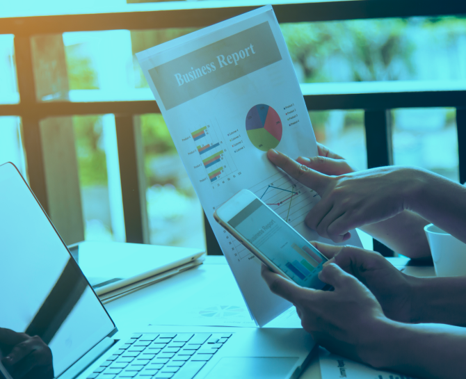 Two people review a business report with graphs, one holding a smartphone and the other pointing to the document, with a laptop nearby.