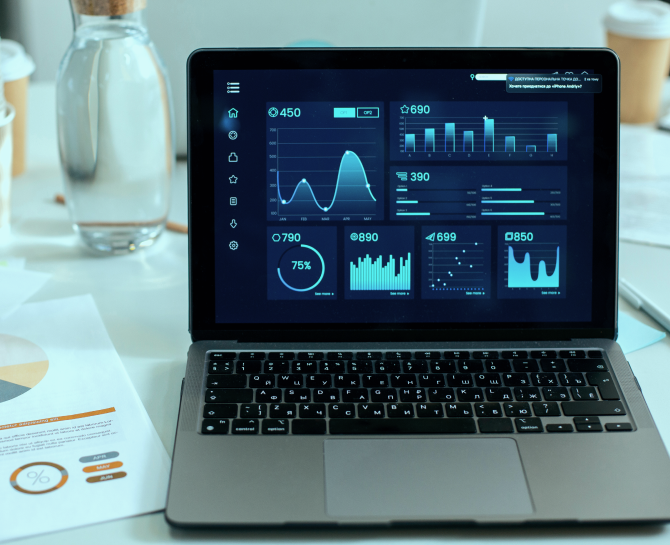 Laptop displaying data analytics dashboard with graphs, charts, and stats. Papers and a water carafe on the table suggest a professional setting.