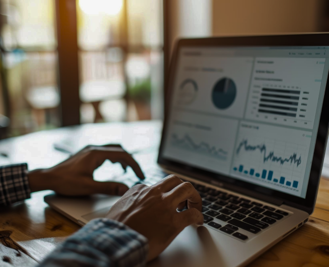 Hands typing on a laptop displaying various charts and graphs, with warm sunlight streaming through distant windows, creating a focused work atmosphere.