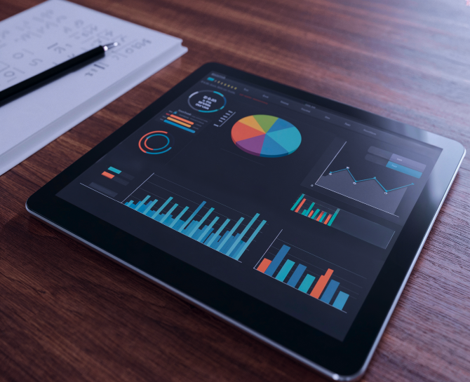 Tablet displaying colorful business charts and graphs on a wooden desk next to a notebook and pen.