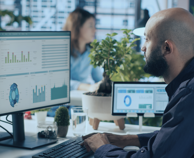 A man in an office analyzes data on a computer, featuring colorful charts and graphs. A plant and another monitor are visible; a woman works in the background.