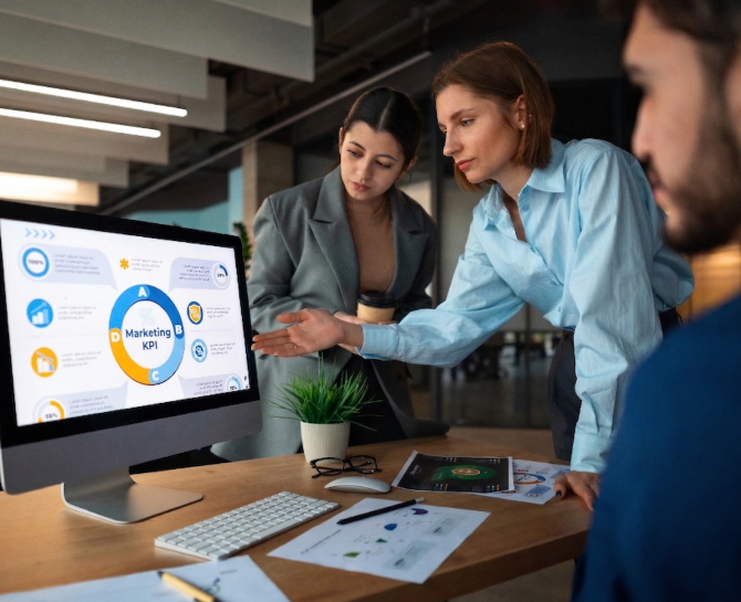 Three colleagues engage in discussion around a computer displaying marketing KPIs. The setting is an office, conveying a focused, collaborative atmosphere.