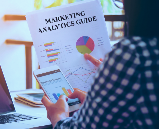 Person analyzing marketing analytics guide and business report with charts on paper and smartphone at desk.