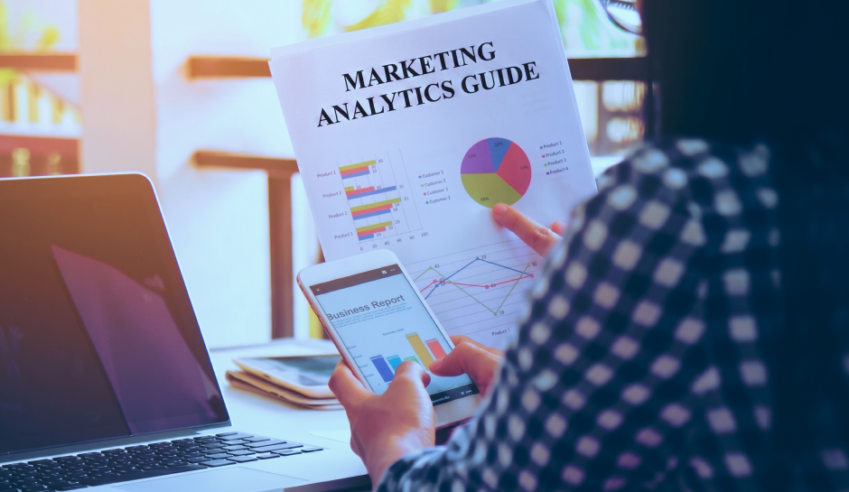 Person analyzing marketing analytics guide and business report with charts on paper and smartphone at desk.